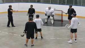 Box lacrosse players in plain uniforms paused near the crease while an official explains positioning.