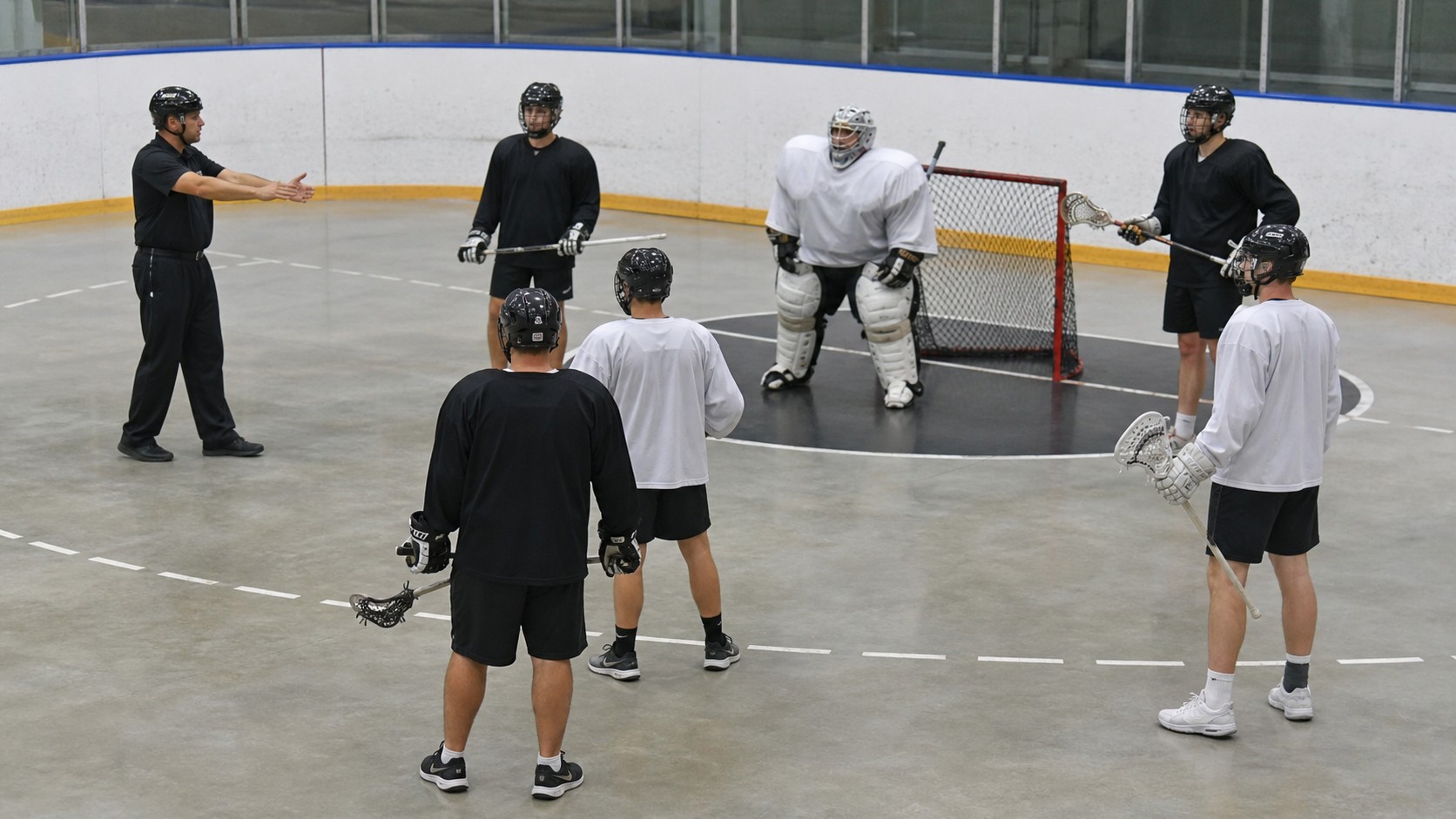 Box lacrosse players in plain uniforms paused near the crease while an official explains positioning.