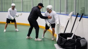 Coach showing a plain-uniform lacrosse player how to use body position near the boards in a box lacrosse rink.