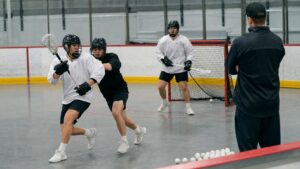 Box lacrosse practice with a plain-uniform player catching under close pressure near the crease.